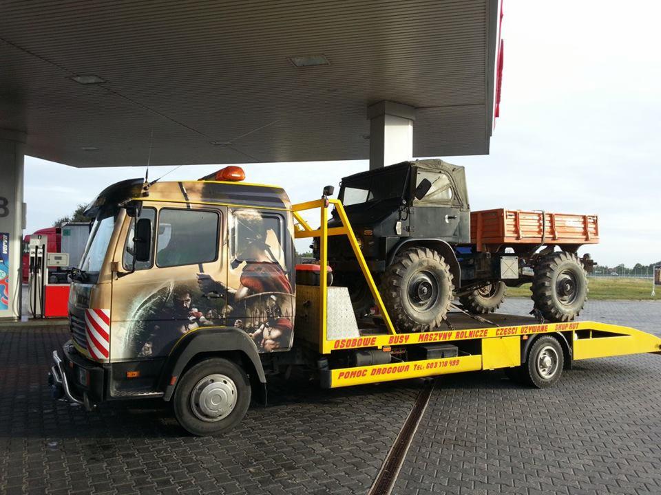 Żółta laweta z grafiką 300 na kabinie, przewożąca Unimoga na stacji benzynowej. Pomoc drogowa z numerem telefonu. Nietypowy wygląd pojazdu.