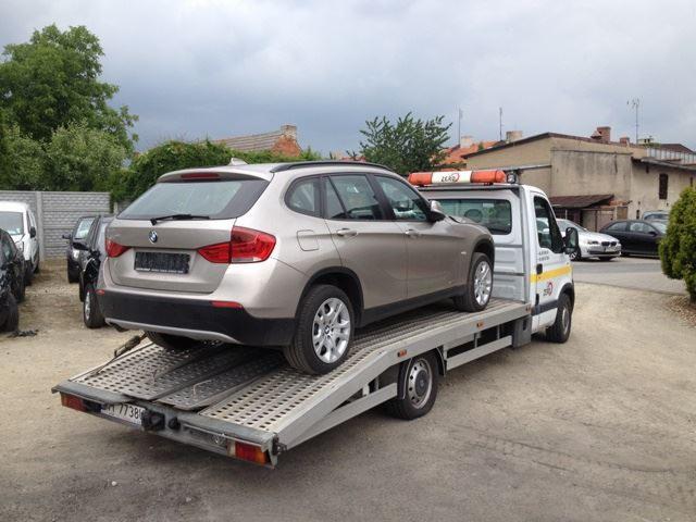 Beżowe BMW X1 na platformie lawety, widok z tyłu. Pomarańczowe światło ostrzegawcze na dachu lawety. Budynki w tle. Tablica rejestracyjna pojazdu widoczna.