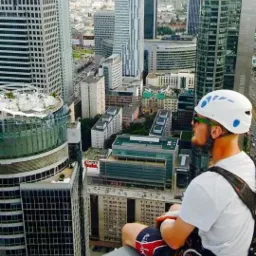 Mężczyzna w kasku siedzi na krawędzi dachu wieżowca, z widokiem na panoramę miasta. Ubrany w uprząż, z linami obok. Lekka mgła w tle.