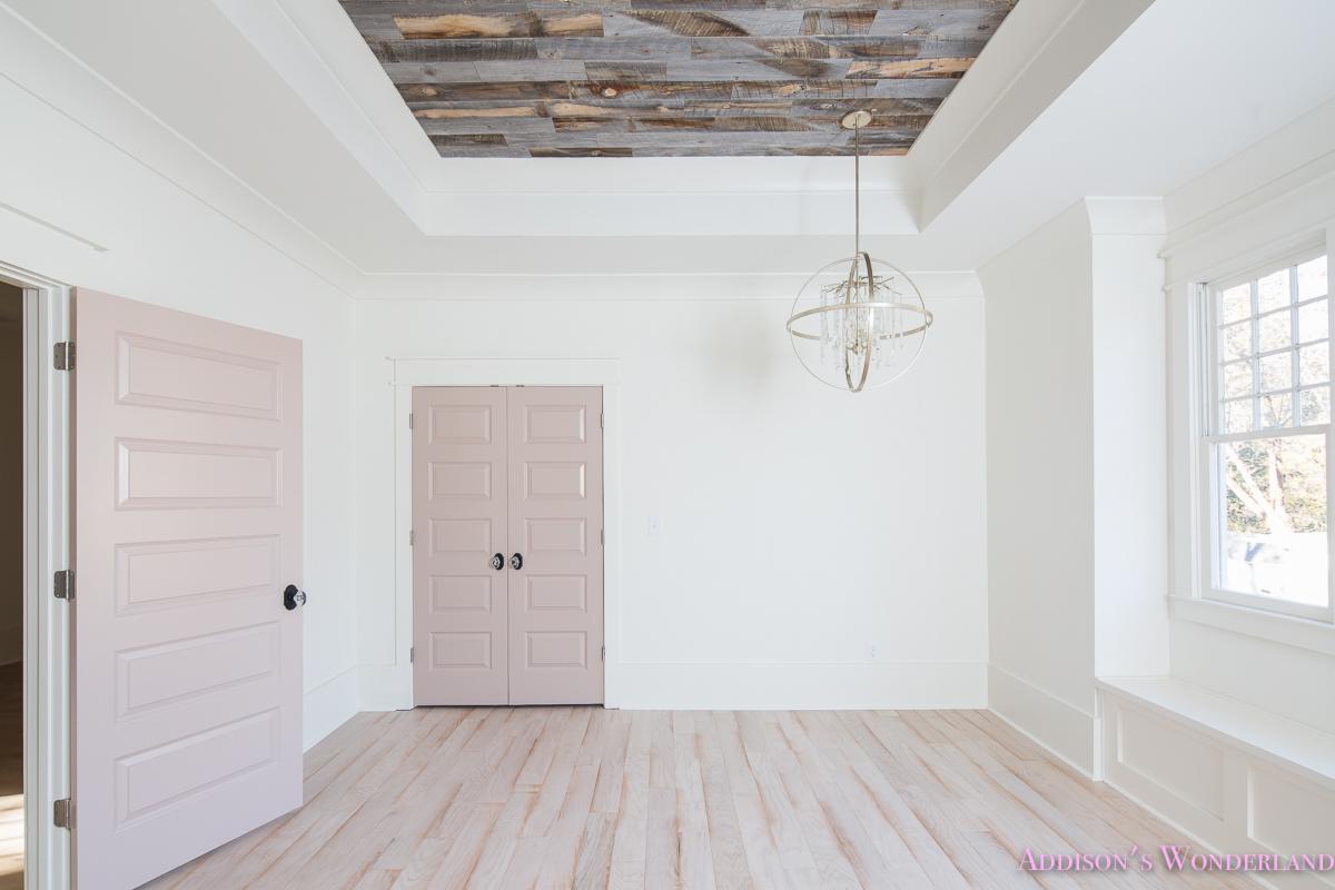 Jasny salon z drewnianym sufitem z recyklingu, parkietem z jasnego drewna, różowymi drzwiami i nowoczesnym żyrandolem kulistym.