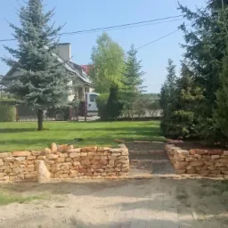 Ogród z kamienną ścieżką i niskim murem oporowym z piaskowca. W tle widoczne drzewa iglaste i trawnik. Częściowo widoczny budynek z jasną elewacją i brązowymi oknami.