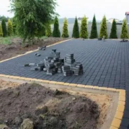 Częściowo ułożona, czarna kostka brukowa z żółtą obwódką. Na powierzchni sterta kostek, w tle rząd drzew iglastych i fragment budynku.