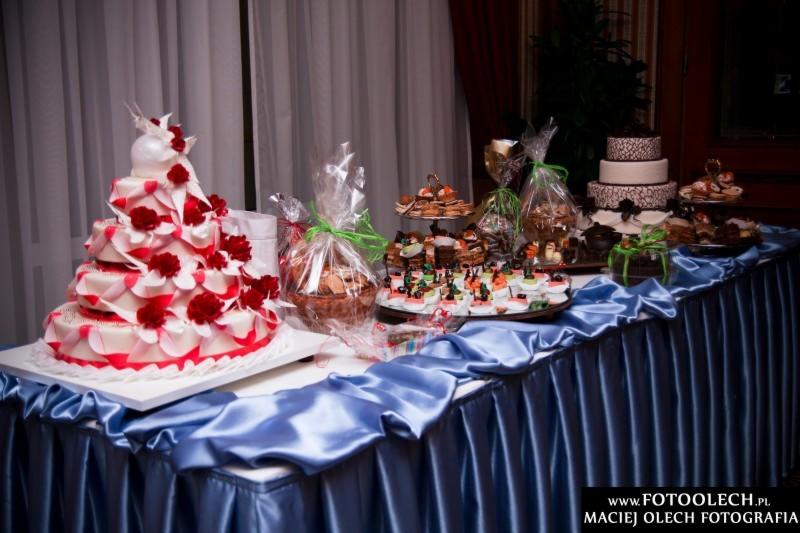 Stół z niebieskim obrusem, na którym znajdują się dwa torty, ciasteczka i prezenty zapakowane w celofan z zielonymi wstążkami. Na pierwszym planie tort piętrowy w kolorze białym z czerwonymi różami.
