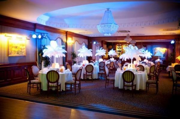 Sala bankietowa z okrągłymi stołami nakrytymi białymi obrusami, dekorowanymi wysokimi, białymi kompozycjami przypominającymi kwiaty lub pióropusze, podwieszony kryształowy żyrandol, drewniane...