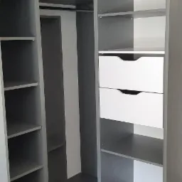 Wnętrze narożnej szafy z przesuwnymi drzwiami, szare półki i białe szuflady, widoczny system prowadnic i drewniana ozdoba sufitowa.