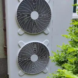 Biała pompa ciepła marki Ariston zamontowana na betonowych wspornikach, widoczna z boku, obok zielonego krzewu i ściany budynku.