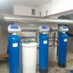 Trzy niebieskie filtry wody Epurotech S1 z białym zbiornikiem na sól w pomieszczeniu z rurami i kafelkami na ścianie.