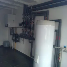 Biały zasobnik ciepłej wody użytkowej z podłączonymi rurami w izolacji termicznej, zamontowany w pomieszczeniu z szarą podłogą i kotłem gazowym na ścianie.