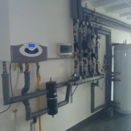 Nowoczesny piec gazowy Ariston z podłączeniami hydraulicznymi, zaworami i manometrami, widoczny zbiornik akumulacyjny, instalacja w pomieszczeniu z szarą podłogą.