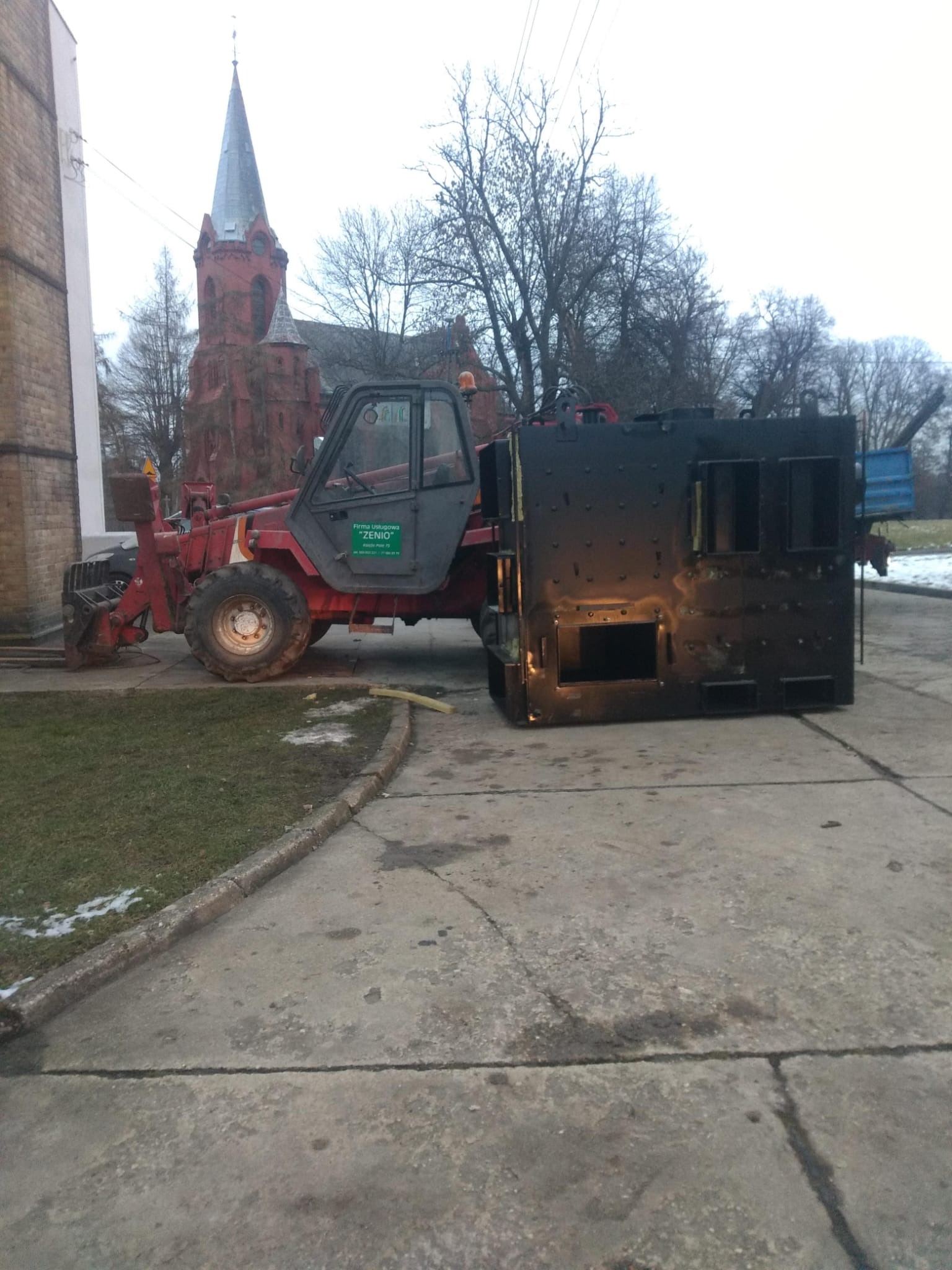 Czerwony wózek widłowy transportuje duży, czarny, metalowy piec na betonowym placu z widocznym kościołem w tle.