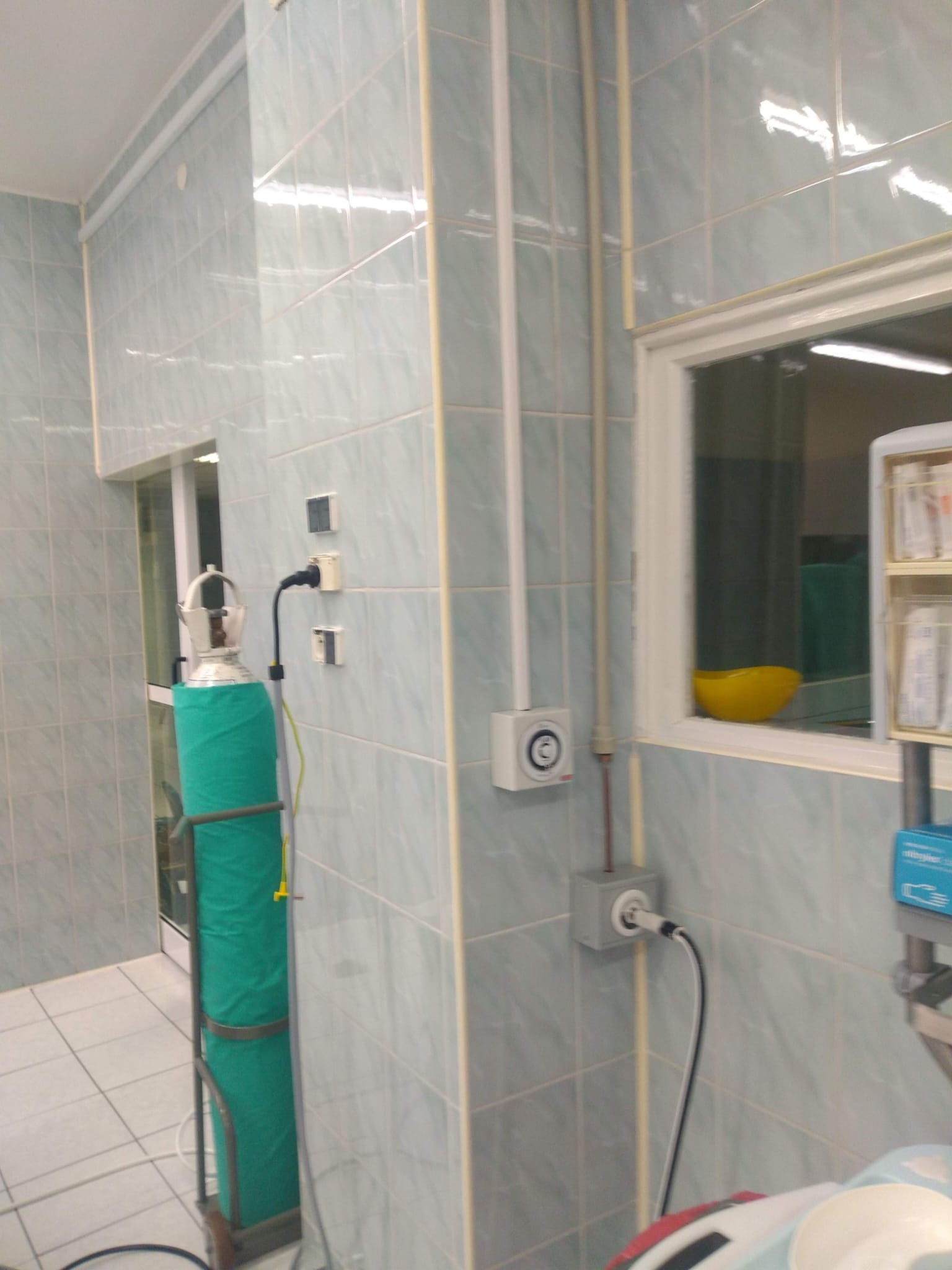 Butla z tlenem w zielonym pokrowcu w pomieszczeniu z błękitnymi kafelkami, widoczne gniazdka elektryczne i okno z żółtą miską w tle.