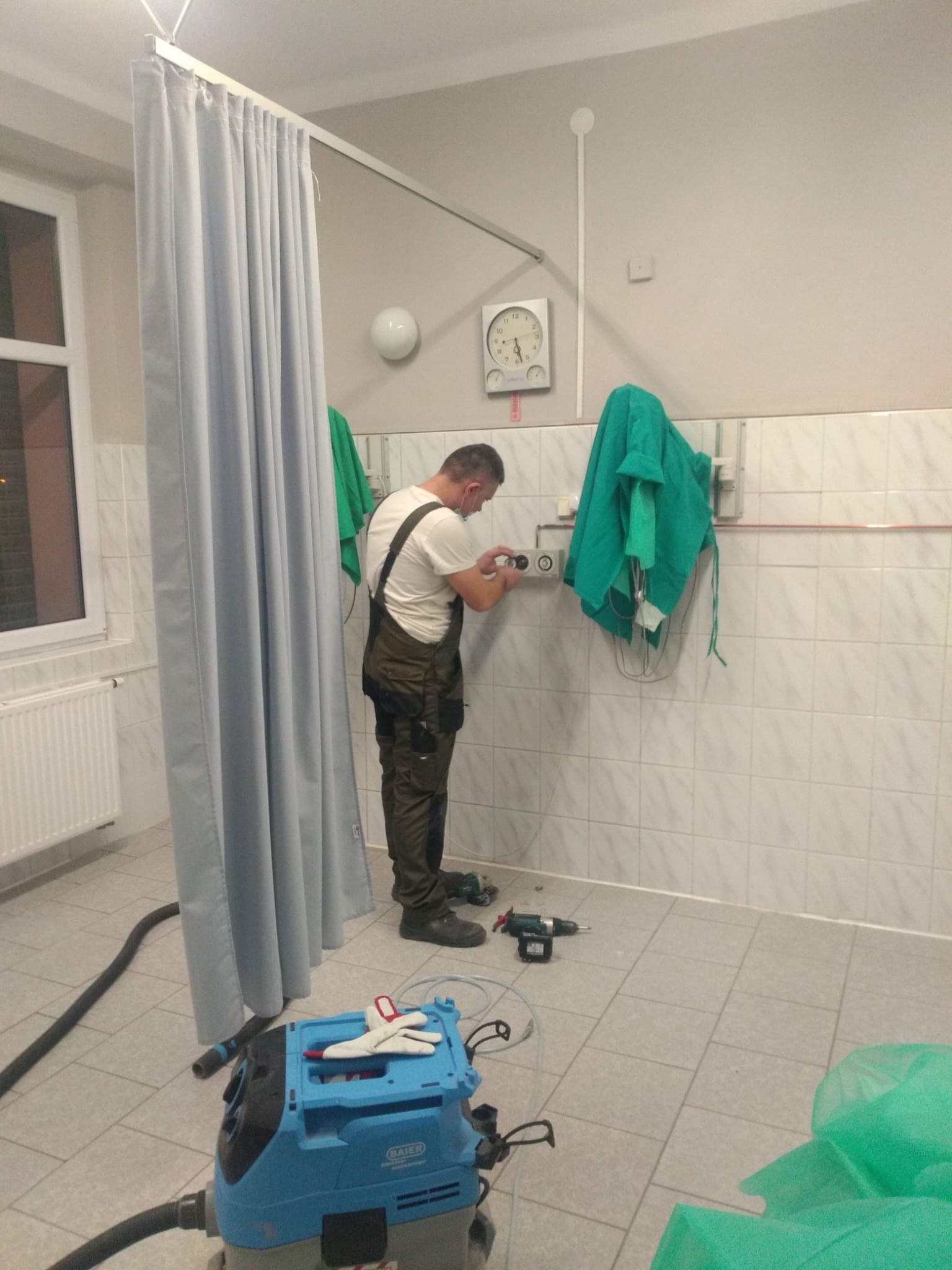 Elektryk w zielonym kombinezonie podłącza gniazdko elektryczne w pomieszczeniu wyłożonym kafelkami, obok leży wiertarka i odkurzacz przemysłowy marki BAIER, wiszą zielone fartuchy medyczne.