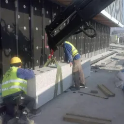 Montaż elementu elewacji z jasnego kamienia na placu budowy. Dźwig podnosi blok, a dwóch robotników w kamizelkach nadzoruje proces. Widoczne narzędzia i materiały budowlane.