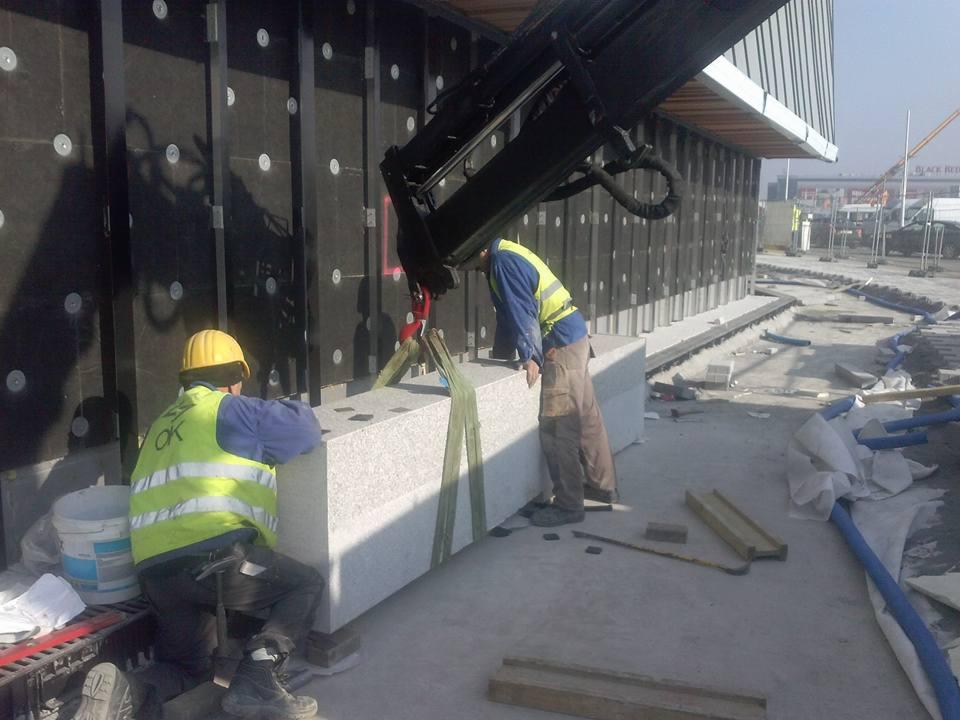 Montaż elementu elewacji z jasnego kamienia na placu budowy. Dźwig podnosi blok, a dwóch robotników w kamizelkach nadzoruje proces. Widoczne narzędzia i materiały budowlane.