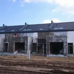 Budowa szeregu domów jednorodzinnych w stanie surowym zamkniętym, widoczne rusztowania, ocieplenie i prace murarskie.