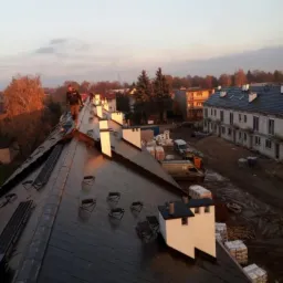 Dekarz spaceruje po dachu z czarnej dachówki, widoczne kominy, zabezpieczenia i budynki w tle o zmierzchu.