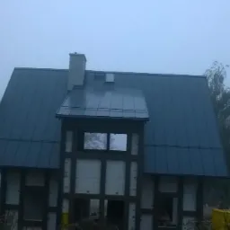 Dom w trakcie budowy z ciemnoszarym, metalowym dachem w mglisty dzień, widoczne elementy konstrukcyjne ścian.