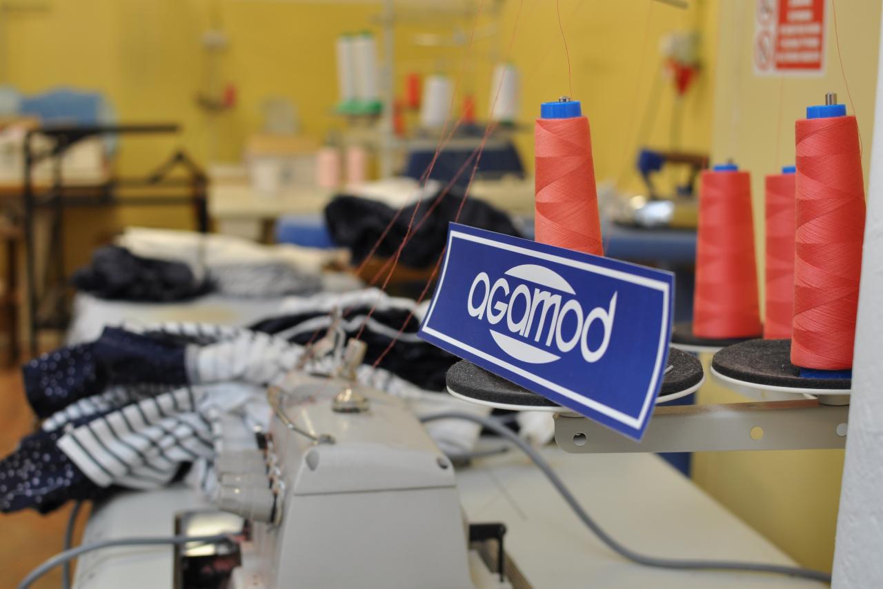 Szwalnia: Detal maszyny do szycia z nawleczoną czerwoną nicią, obok leżą materiały w paski i kropki, a w tle widoczne stanowiska pracy i nici.