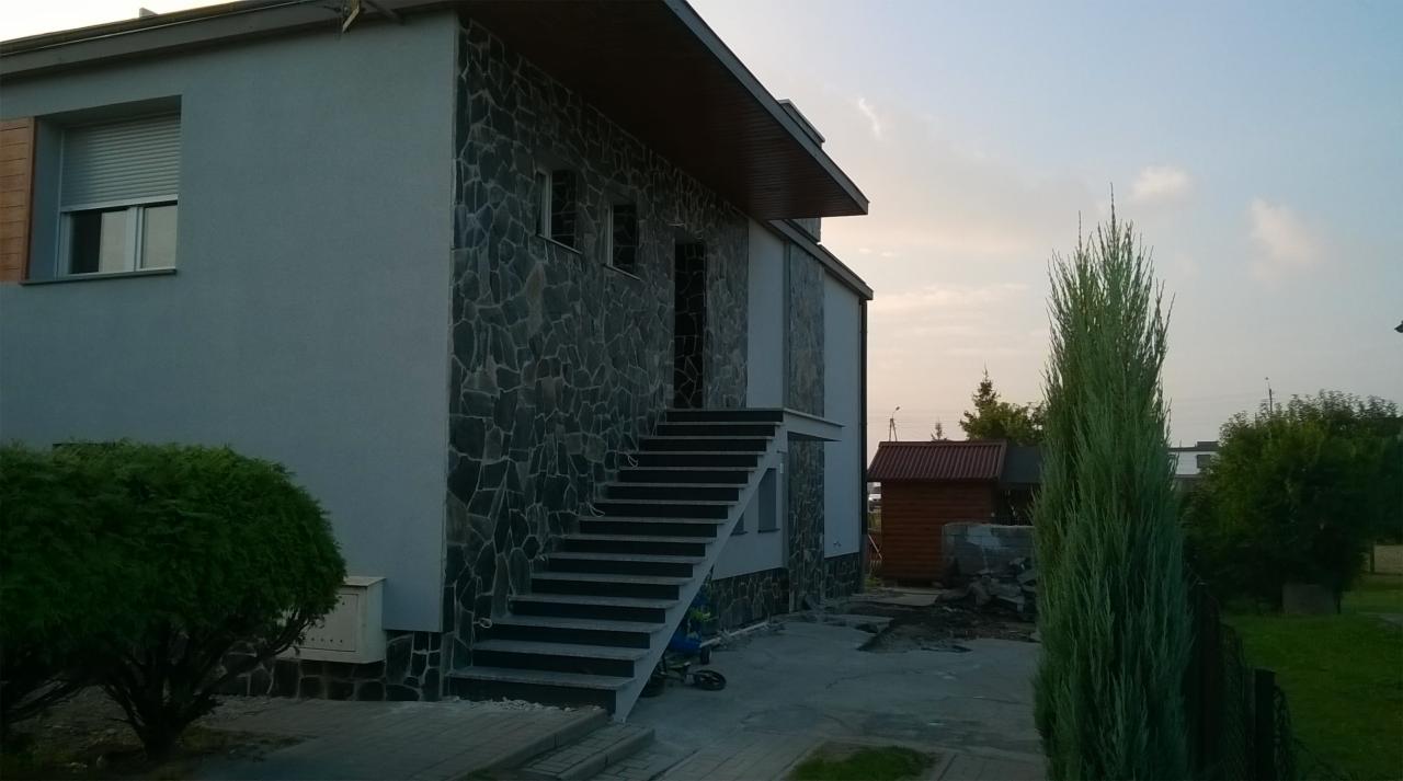 Wykończona elewacja domu z szarego tynku i fragmentami okładziny z ciemnego kamienia łupanego, schody zewnętrzne prowadzące do wejścia na piętro, widok z boku w pochmurny dzień.