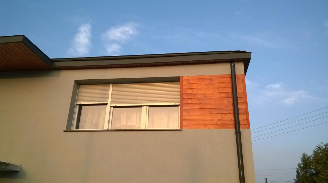 Fragment elewacji budynku z szarą fasadą, drewnianą okładziną i oknem z opuszczonymi roletami, widoczny również element orynnowania w kolorze czarnym.