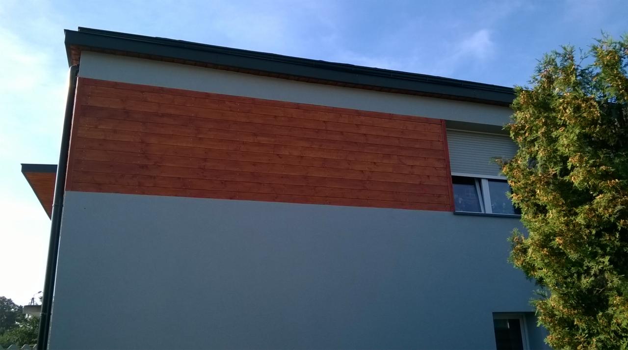 Fragment elewacji budynku z poziomym pasem z desek z drewna egzotycznego, szary tynk, okno z roletą zewnętrzną i fragment iglastego drzewa.