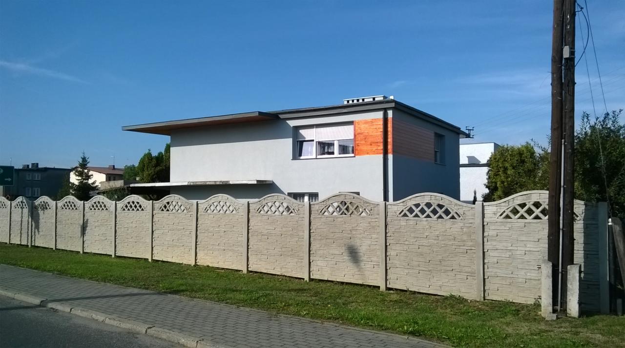 Nowoczesny dom jednorodzinny z szarą elewacją, drewnianymi akcentami i ogrodzeniem z prefabrykatów betonowych o dekoracyjnym wzorze.