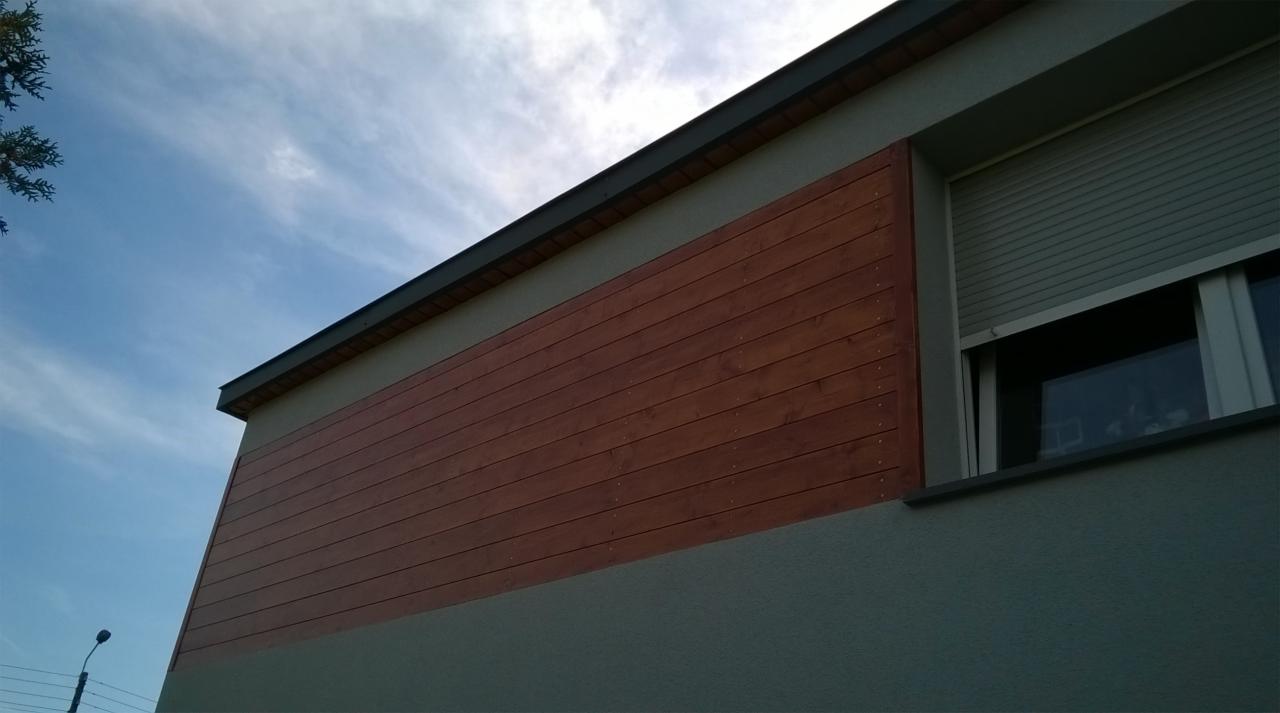 Fragment elewacji budynku z drewnianą okładziną w kolorze brązowym i szarą fasadą, widoczny fragment okna z roletą zewnętrzną oraz błękitne niebo.