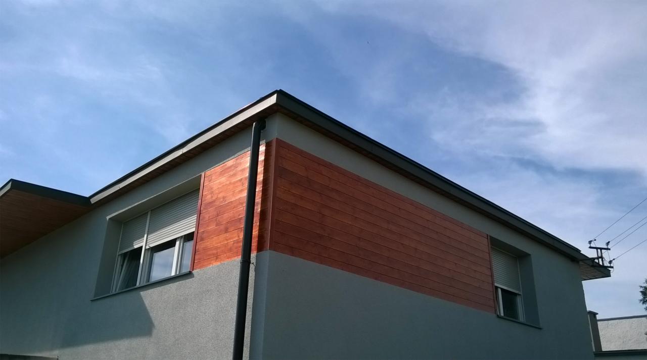 Fragment elewacji domu z wyraźnym podziałem na szary tynk i poziome deski z drewna egzotycznego, widoczne okno z roletą, rynna i błękitne niebo.