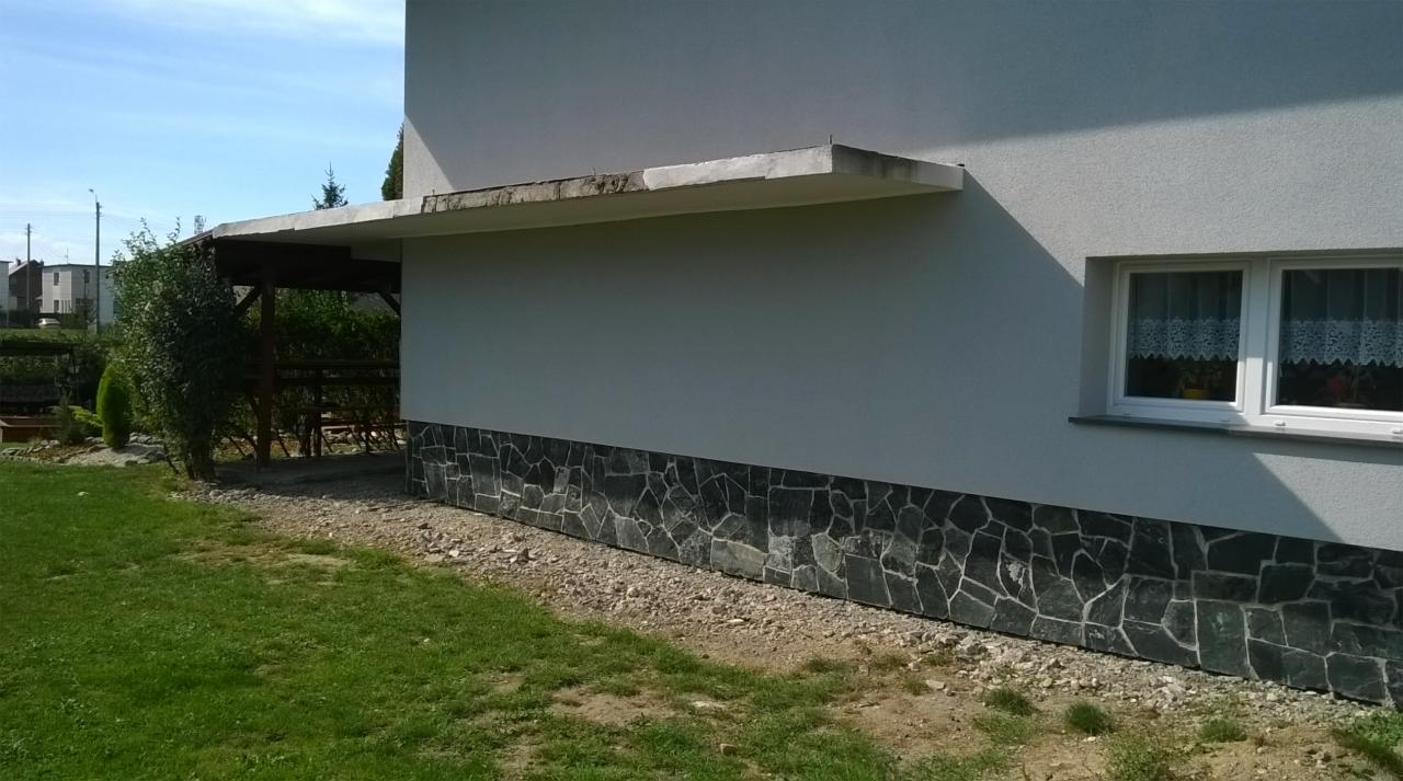 Elewacja budynku w Krzanowicach z szarą fakturą i kamienną podmurówką z nieregularnych, ciemnych kamieni. Widoczny fragment ogrodu z zadaszeniem.