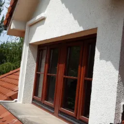 Widok z zewnątrz na okno dachowe z brązową ramą, zamontowane w białej ścianie budynku w Niemczy, z widocznym fragmentem dachu pokrytego czerwoną dachówką i zielenią drzew w tle.