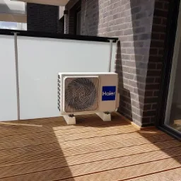 Jednostka zewnętrzna klimatyzacji Haier 3.5kW zamontowana na drewnianym tarasie balkonu, widok z boku, szara cegła elewacyjna w tle.