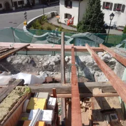 Demontaż starego dachu z widoczną konstrukcją więźby dachowej i zabezpieczeniami siatkowymi, w tle budynki mieszkalne i szwajcarska flaga.