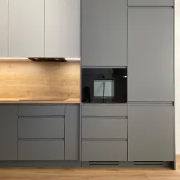 Nowoczesna zabudowa kuchenna w odcieniach szarości i bieli, z drewnianym blatem i czarnym piekarnikiem wbudowanym w szafkę, oświetlenie LED pod szafkami górnymi, widoczna podłoga z paneli...