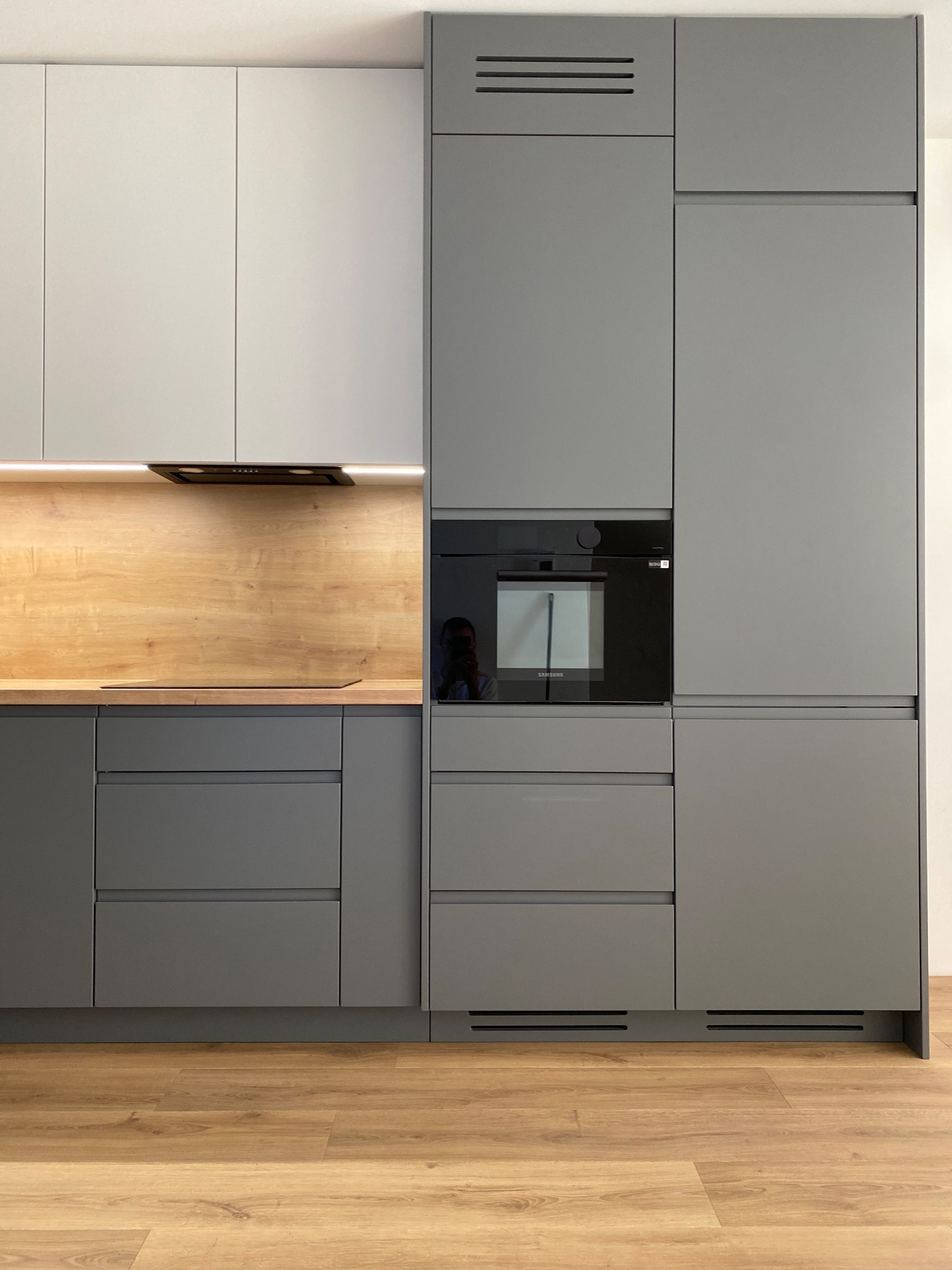 Nowoczesna zabudowa kuchenna w odcieniach szarości i bieli, z drewnianym blatem i czarnym piekarnikiem wbudowanym w szafkę, oświetlenie LED pod szafkami górnymi, widoczna podłoga z paneli...