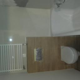 Remonty i wykończenia wnętrz TEAM CONCEPT - Jasna łazienka po remoncie: biały grzejnik, brązowe płytki imitujące drewno, biała toaleta i wanna. Widoczny fragment sufitu z niedoskonałościami.