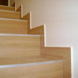 Remonty i wykończenia wnętrz TEAM CONCEPT - Schody wykończone panelami imitującymi drewno, z aluminiowymi listwami na krawędziach, przylegające do jasnej ściany. Detal wykończenia wnętrza.