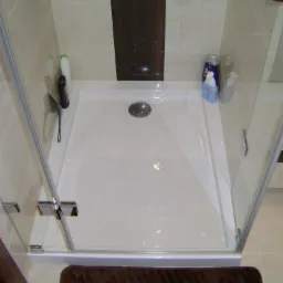 Nowoczesna kabina prysznicowa z brodzikiem i szklanymi drzwiami, wykończona beżowymi płytkami. Na brodziku widoczne kosmetyki, a na podłodze brązowy dywanik.