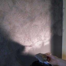 Remonty i wykończenia wnętrz TEAM CONCEPT - Nakładanie dekoracyjnej, szarej gładzi strukturalnej na ścianę za pomocą pędzla i wiadra. Widoczne nieregularne wzory i faktura na powierzchni.