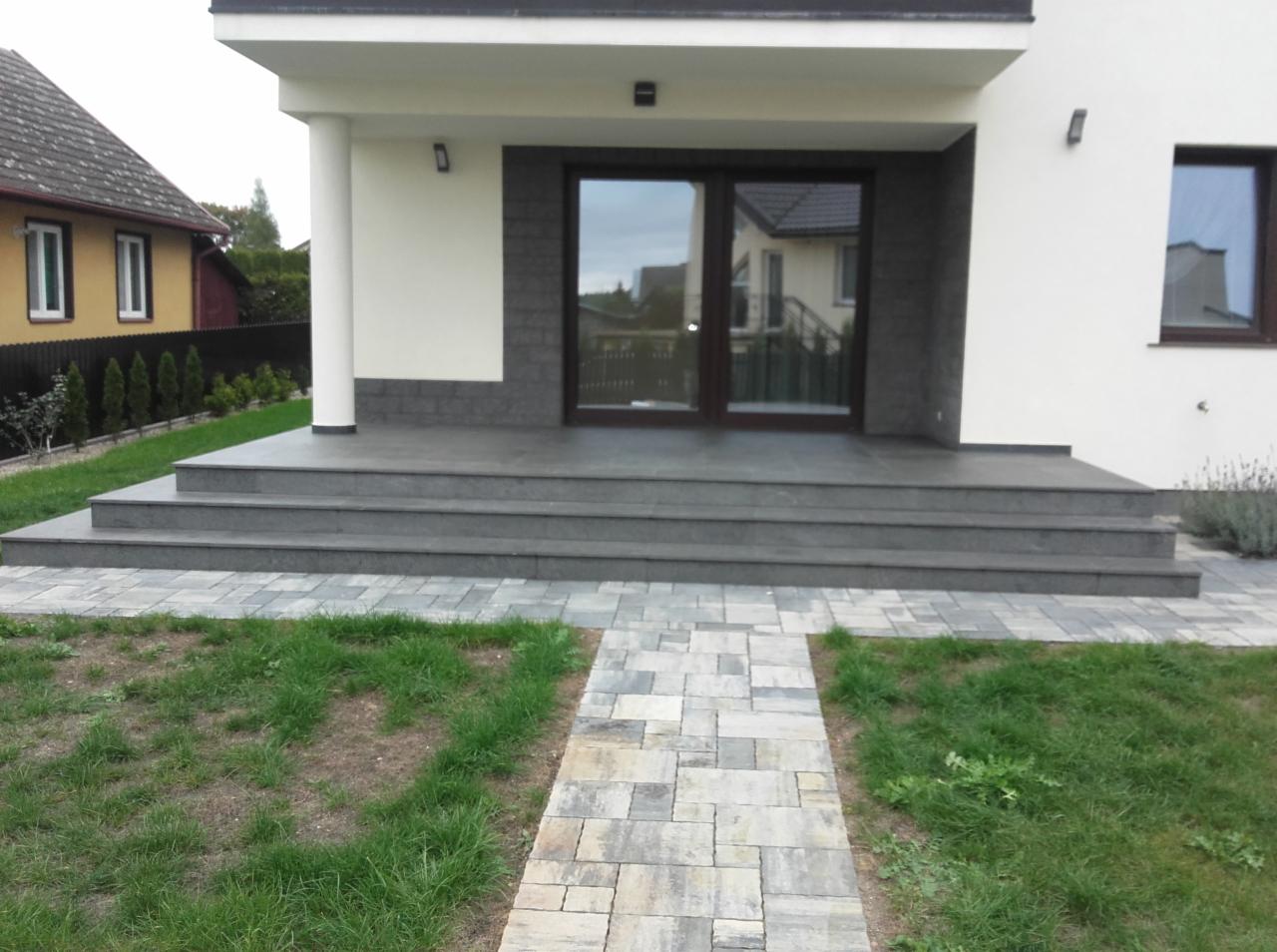 Nowoczesne wykończenie wejścia do domu z szarymi płytkami na schodach i ścieżką z kostki brukowej. Widoczna kolumna i okno.