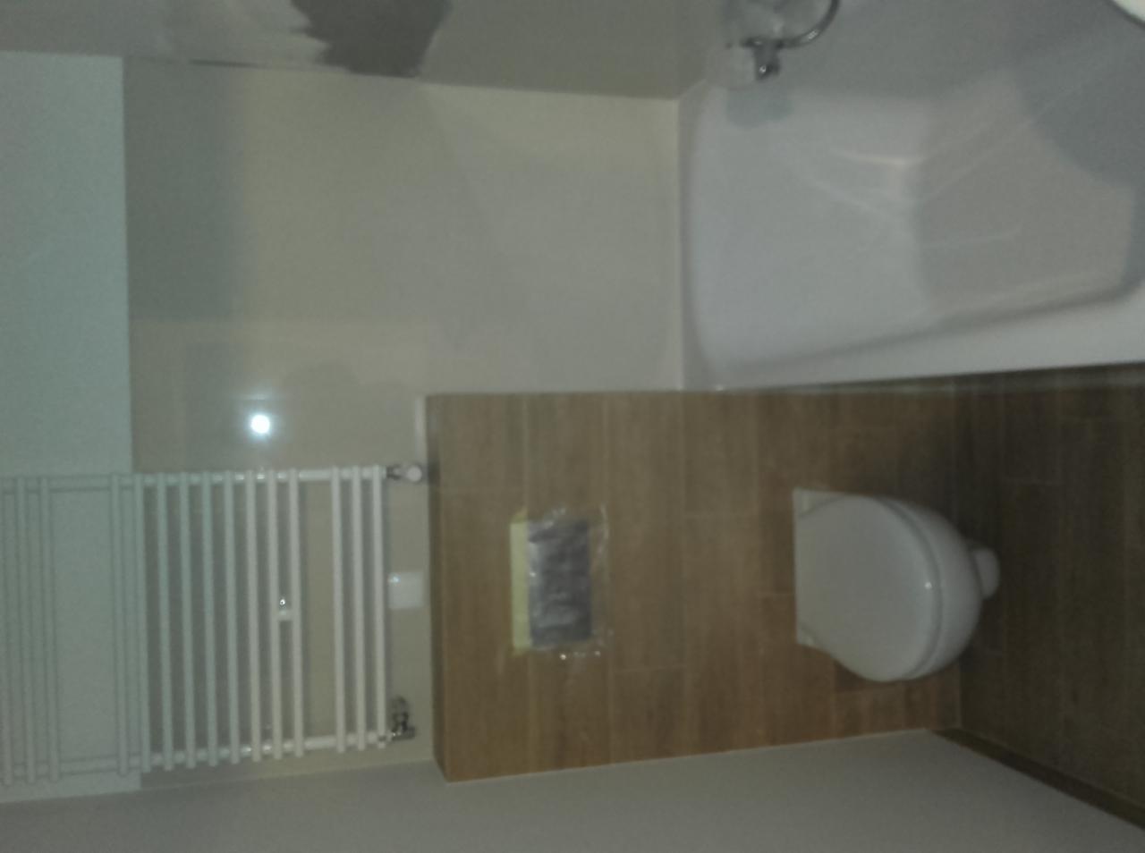 Jasna łazienka po remoncie: biały grzejnik, brązowe płytki imitujące drewno, biała toaleta i wanna. Widoczny fragment sufitu z niedoskonałościami.