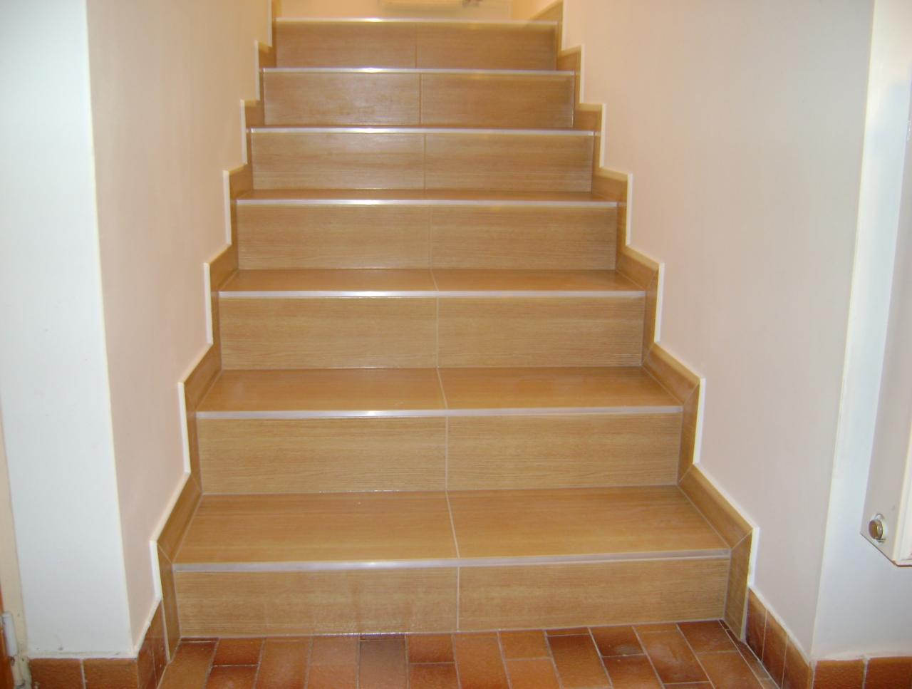 Schody wyłożone płytkami imitującymi drewno, z listwami wykończeniowymi. Widoczne brązowe płytki podłogowe i białe ściany. Kąt ujęcia od dołu.