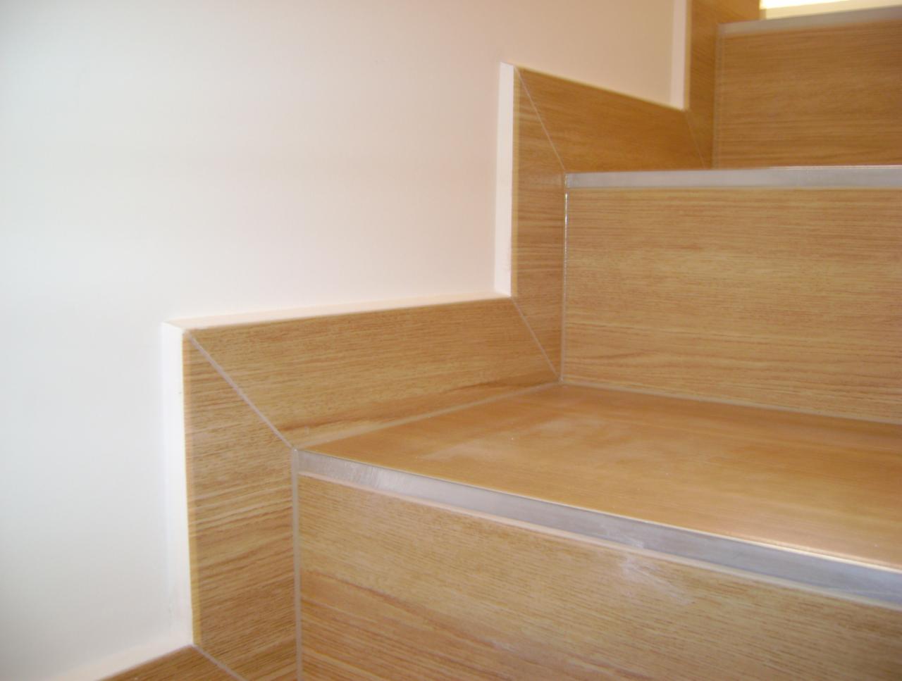 Wykończone schody z płytek imitujących drewno, z listwami zabezpieczającymi krawędzie. Biała ściana w tle, minimalistyczne detale wykończenia.