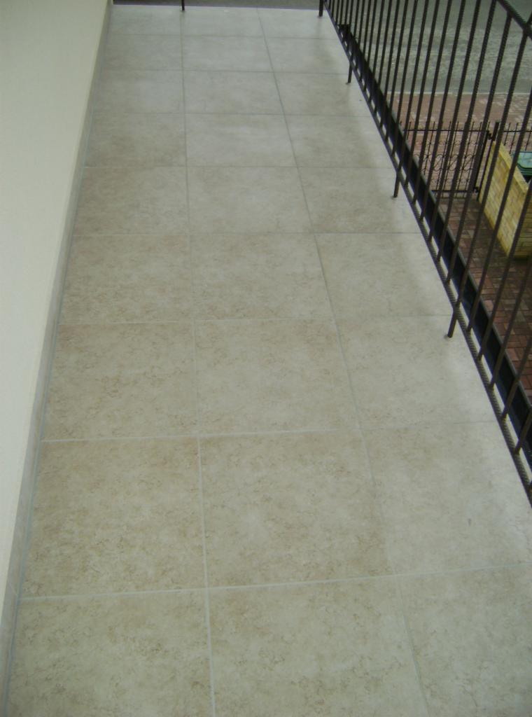 Beżowe płytki podłogowe na balkonie z czarną, metalową barierką. Widok z góry, ukazujący regularny wzór ułożenia płytek i fragment otoczenia za barierką.