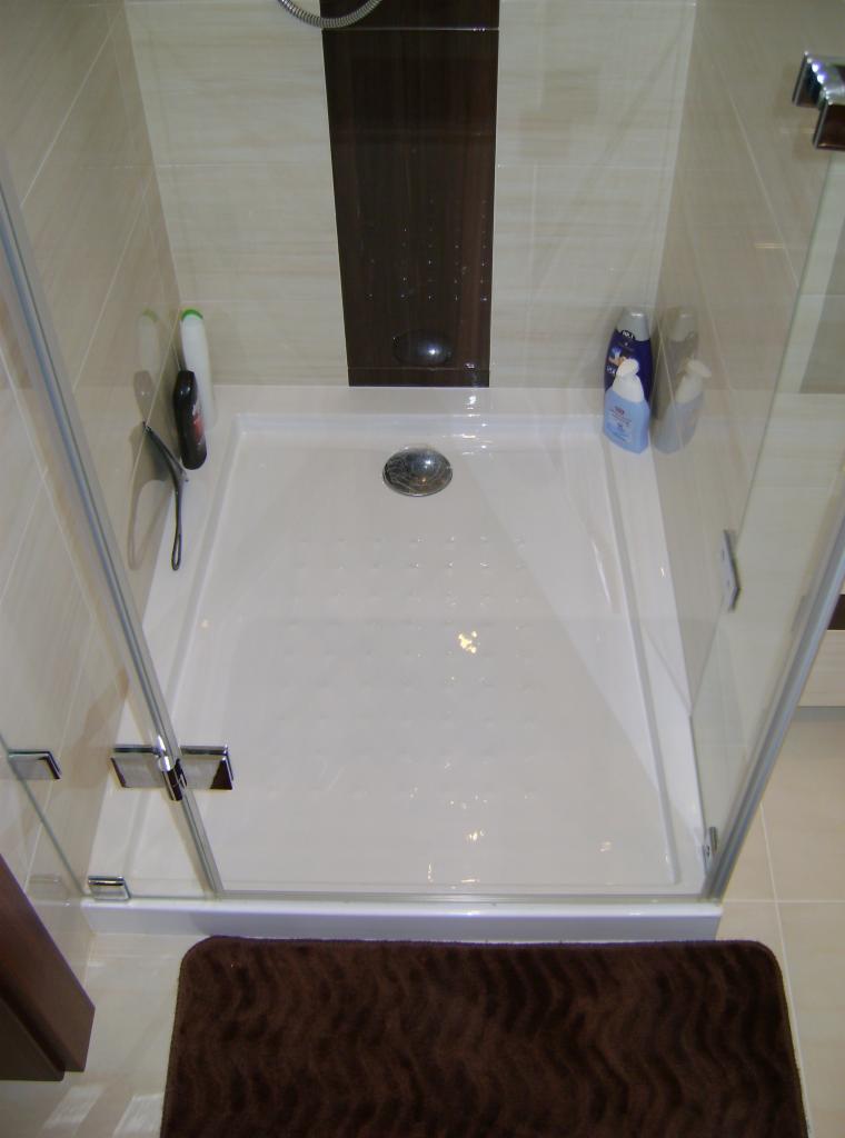 Nowoczesna kabina prysznicowa z brodzikiem i szklanymi drzwiami, wykończona beżowymi płytkami. Na brodziku widoczne kosmetyki, a na podłodze brązowy dywanik.