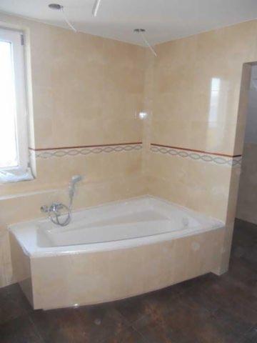 Wykończona łazienka z wanną obudowaną płytkami w odcieniach beżu, z dekoracyjnym pasem płytek i drewnopodobną podłogą. Widoczne wyprowadzone kable.