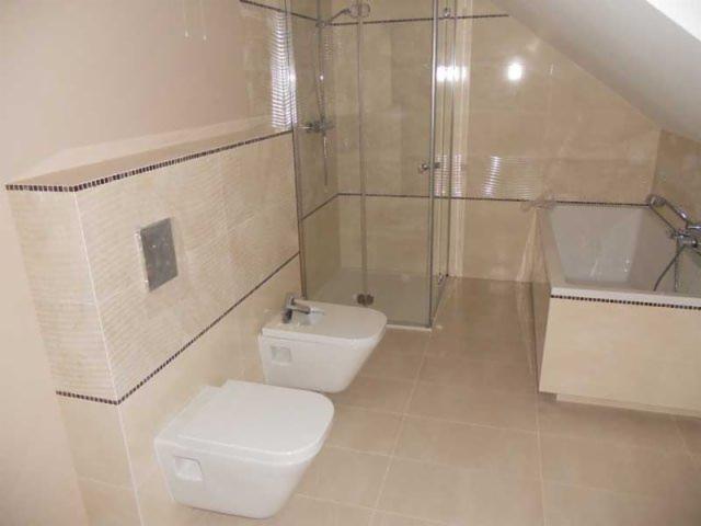 Beżowa łazienka na poddaszu ze szklaną kabiną prysznicową, wanną i podwieszaną toaletą z bidetem. Wykończenie płytkami z czarnym akcentem.