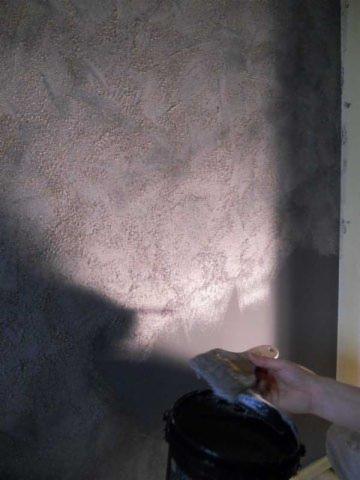 Nakładanie dekoracyjnej, szarej gładzi strukturalnej na ścianę za pomocą pędzla i wiadra. Widoczne nieregularne wzory i faktura na powierzchni.