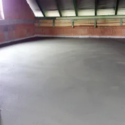 Świeżo wylana, gładka, szara posadzka w surowym wnętrzu poddasza z ceglanymi ścianami i drewnianą konstrukcją dachu.