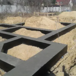 Zalane fundamenty domu z izolacją pionową, wypełnione piaskiem, z wystającymi prętami zbrojeniowymi. Widoczny wykop i drewniane znaczniki.