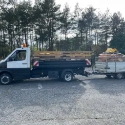 Ciężarówka z przyczepą wypełniona drewnem na tle drzew, widok z boku na szarym asfalcie.